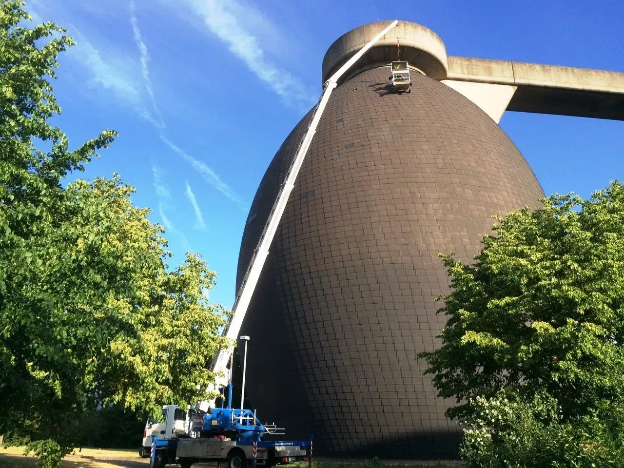 Ein großes braunes Silo unter blauem Himmel, umgeben von einem Kran und Bäumen.