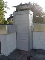 Weißer Dachschornstein mit Metallabdeckung zwischen niedrigen Stuckwänden unter Bäumen