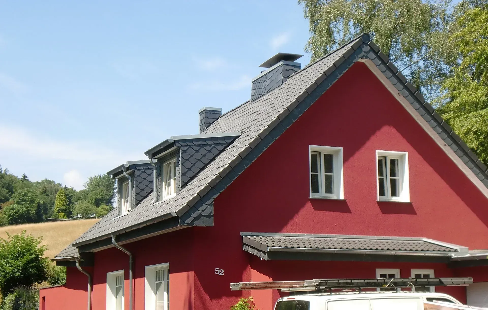 Rotes Haus mit dunklem Dach und Dachgauben unter blauem Himmel, teilweise verdeckt von Bäumen und einem geparkten Fahrzeug