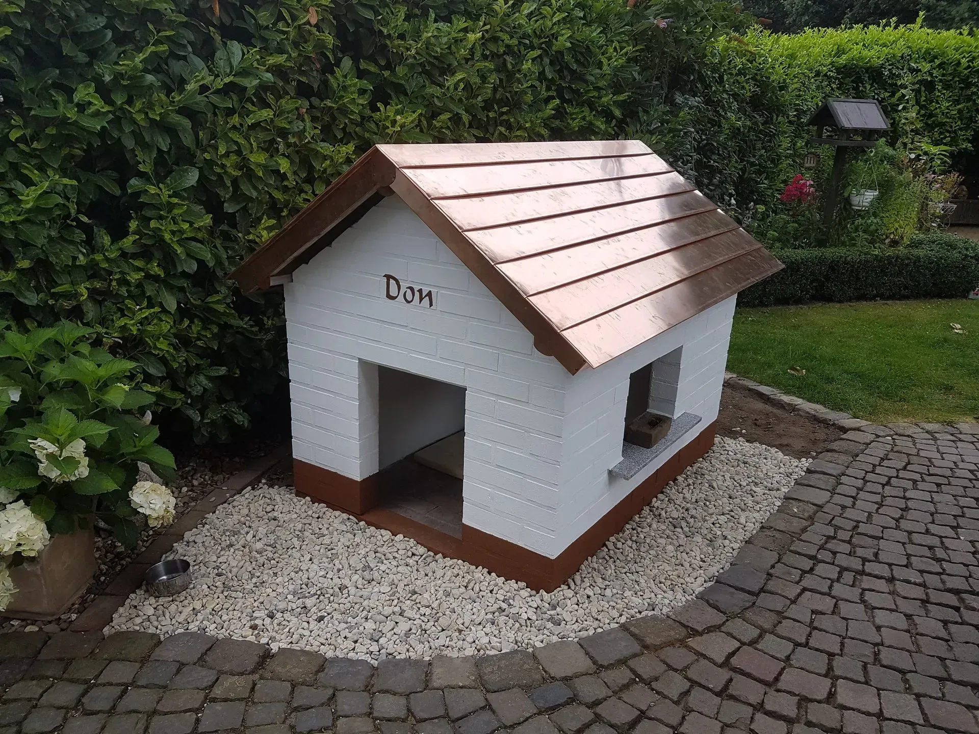 Kleine weiße Hundehütte mit Kupferdach auf einem Kiesplatz neben einem Backsteinweg und Gartengrün