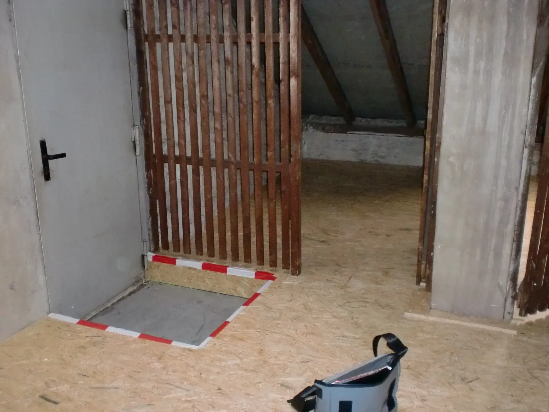 Kleiner, unfertiger Raum mit einem Holztor, offener Tür und einer schwarzen Tasche auf dem Boden.