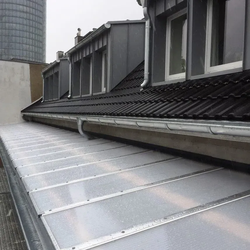 Blick vom Dach eines grauen Dachgaubengebäudes mit Metalldachplatten und Dachrinnen bei bewölktem Licht