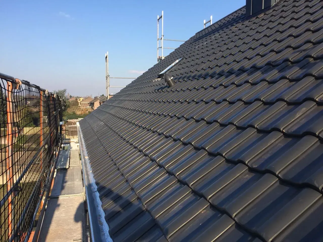 Dunkles Ziegeldach mit Solarpaneelen und daneben ein Sicherheitszaun unter klarem Himmel