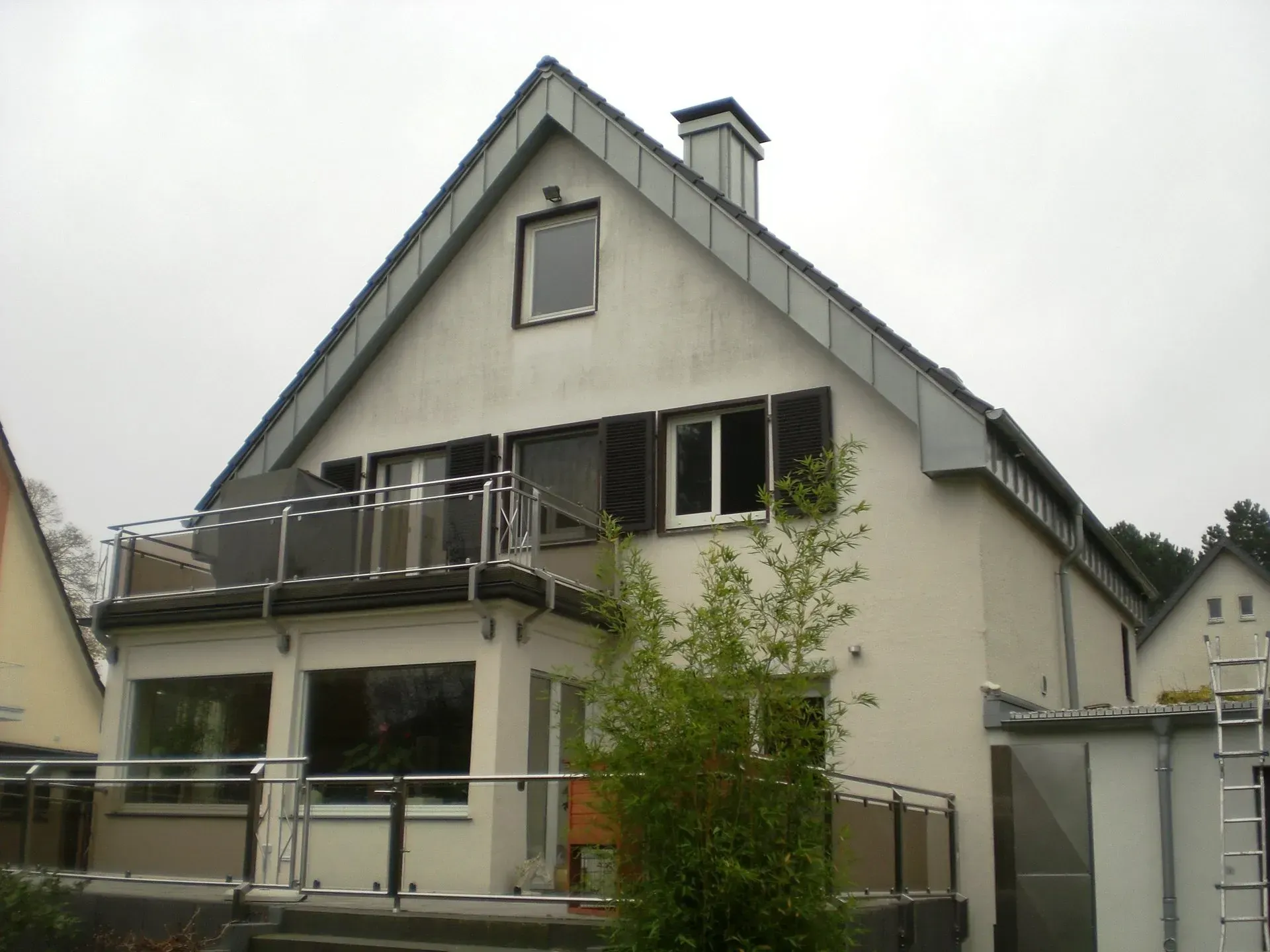 Zweistöckiges weißes Haus mit Balkon, dunklem Dach und kleinen Bäumen davor