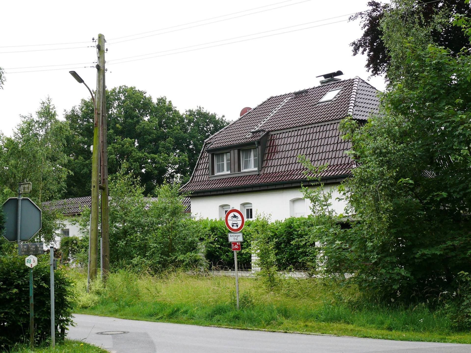 Ländliches Haus am Straßenrand mit rotem Ziegeldach, Bäumen und einem Verkehrsschild an einer kurvigen Straße