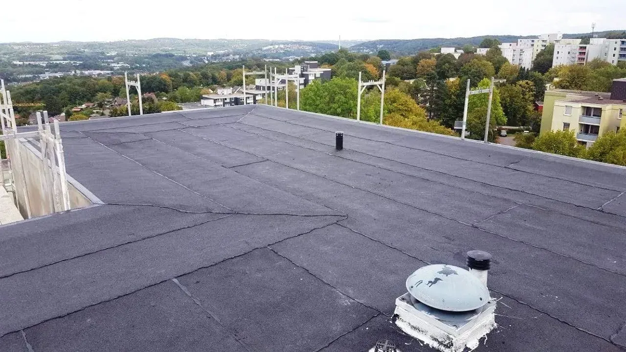 Flaches, schwarzes Dach mit Lüftungsschlitzen, das unter klarem Himmel auf eine ferne Stadtlandschaft blickt.