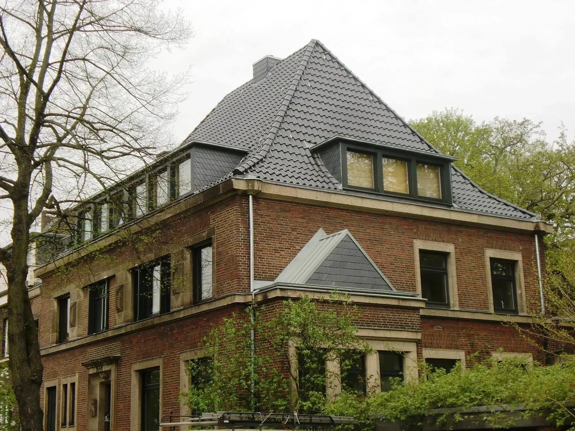 Backsteinhaus mit dunklem Dach und Dachgauben, von der Straße aus inmitten von Bäumen und Grünflächen gesehen