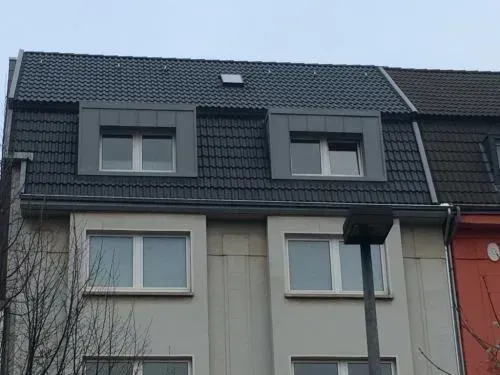 Fassade eines Wohngebäudes mit dunklen Dachziegeln, grauen Wänden und einem roten Abschnitt auf der rechten Seite