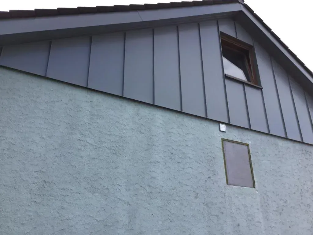 Graue Hausfassade mit dunklem Dach und zwei kleinen Fenstern unter einem bewölkten Himmel