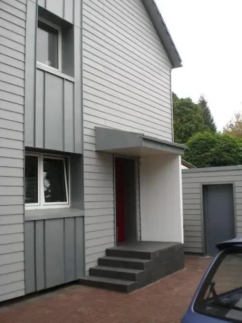 Moderner grauer Hauseingang mit Treppe, Haustür und einer kleinen freistehenden Garage neben einer gepflasterten Auffahrt.