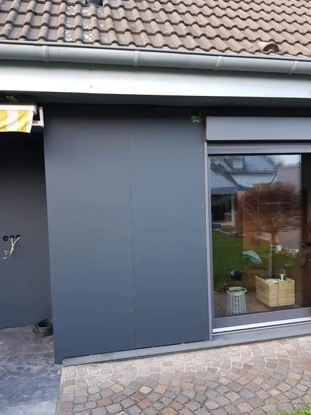 Dunkelgraue Ladenfront mit großem Fenster, Ziegeldach und Kopfsteinpflaster-Gehweg.