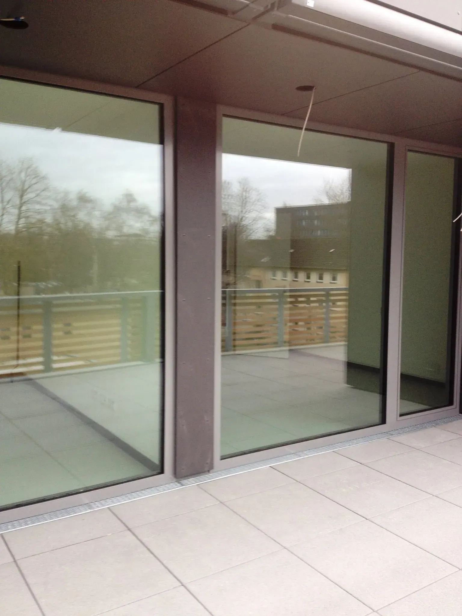 Leerer Balkon mit großen Glasschiebetüren und Fliesenboden, mit Blick auf ein Holzgeländer und kahle Bäume.