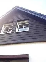 Hausgiebel mit dunkler Verkleidung und zwei weiß gerahmten Fenstern über einer weißen Wand