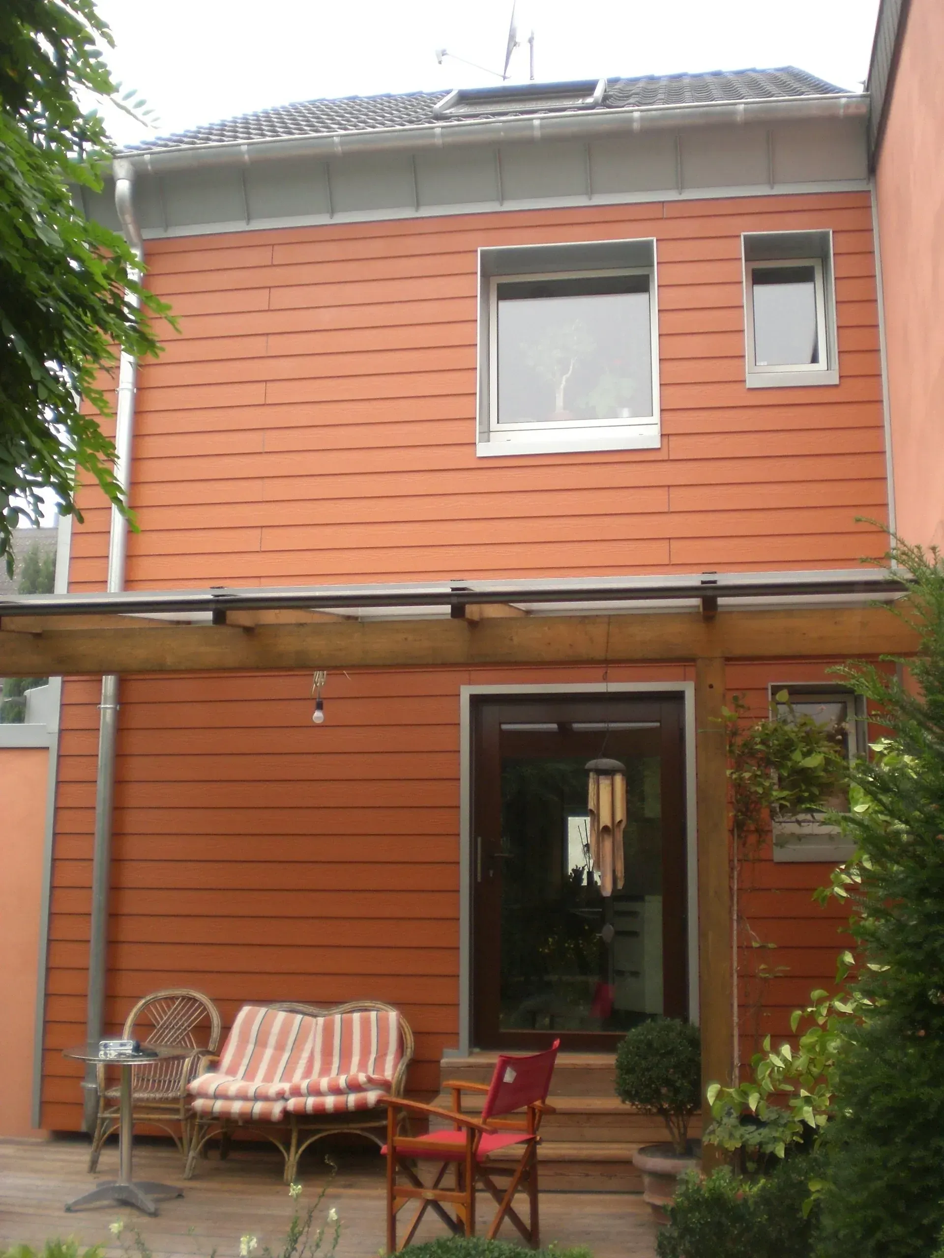 Zweistöckiges orangefarbenes Haus mit kleiner Veranda, Terrassenstühlen und viel Grün im Außenbereich.