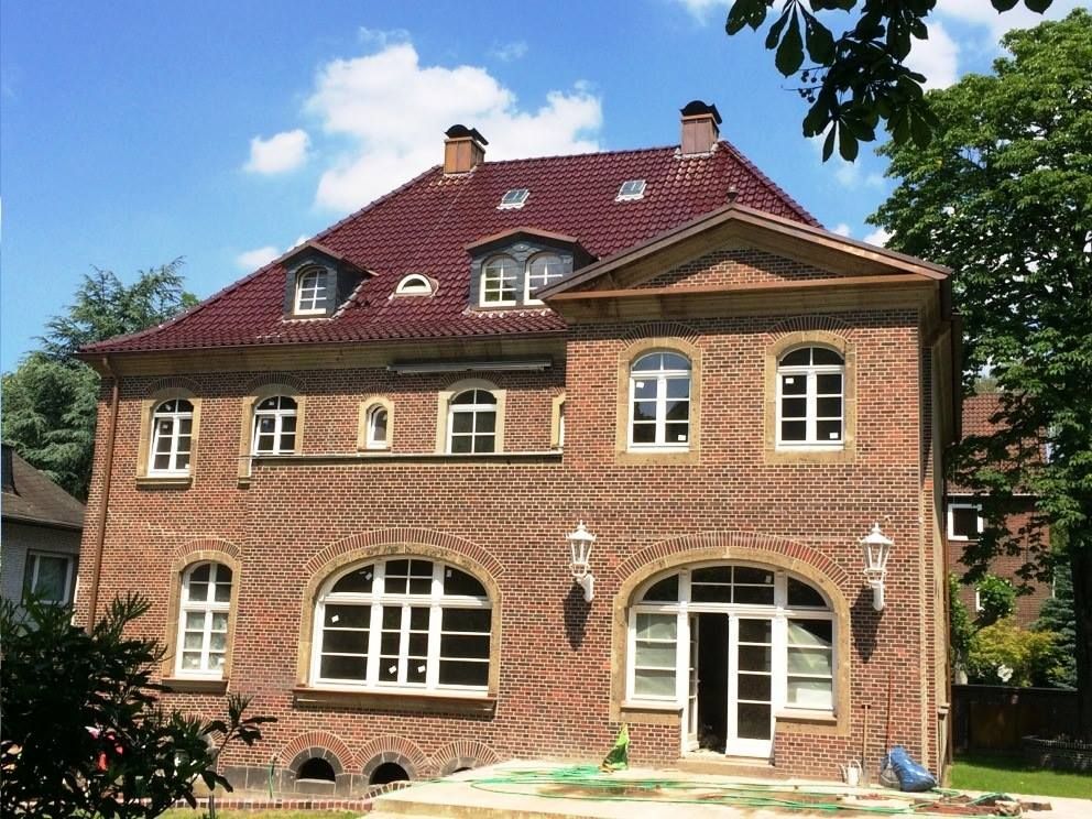 Großes Backsteinhaus mit rotem Dach, weiß umrandeten Fenstern und Bogentüren unter belaubten Bäumen