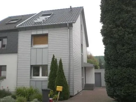 Graues Haus mit Solarpaneelen auf dem Dach, einem kleinen Vorgarten und einer Auffahrt neben hohen Hecken.