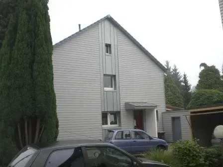Graues Haus mit steilem Dach, geparkte Autos und hohe Bäume in einer Wohnauffahrt.