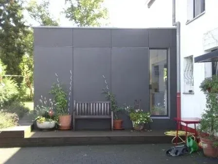 Kleiner grauer Gartenschuppen mit Bank und Topfpflanzen neben einem Haus