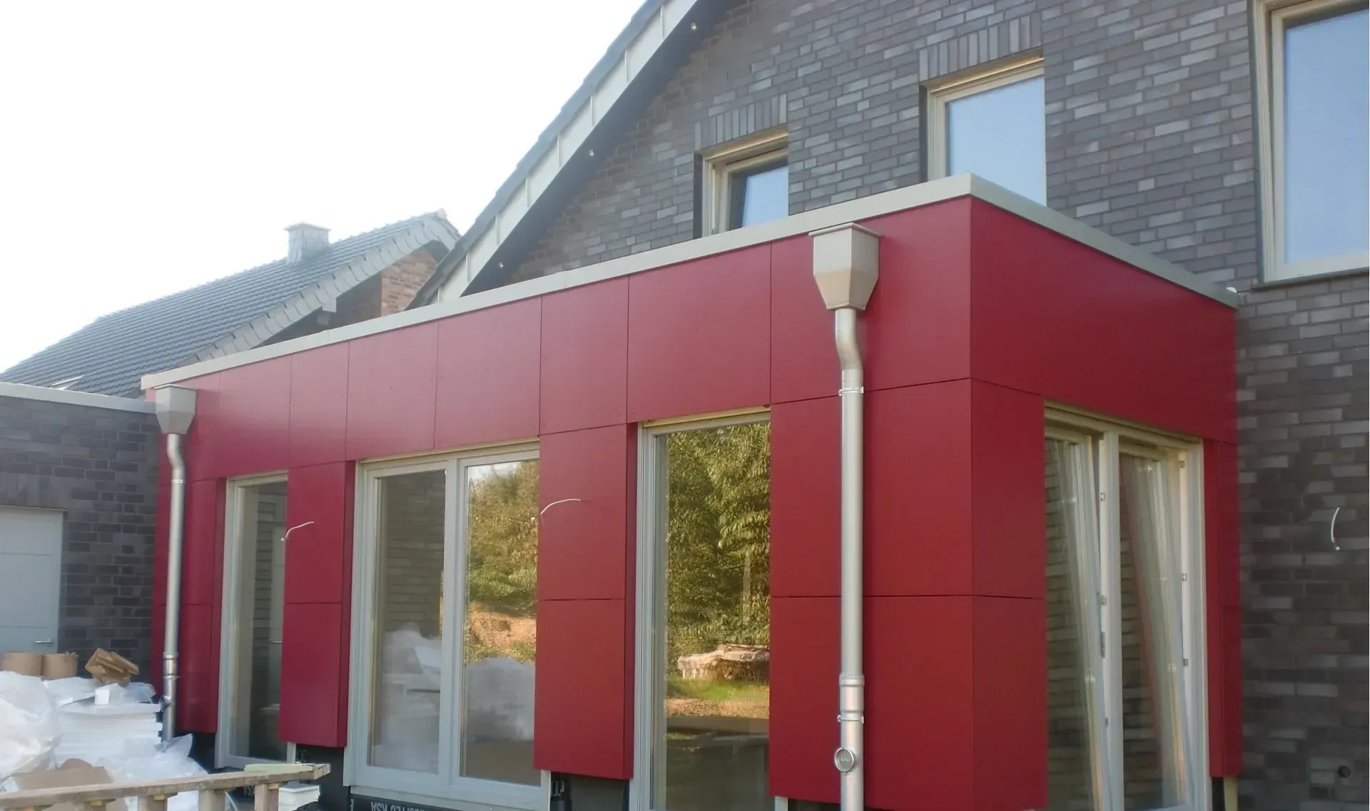 Moderner roter Hausanbau mit großen Fenstern an ein im Bau befindliches graues Backsteinhaus.