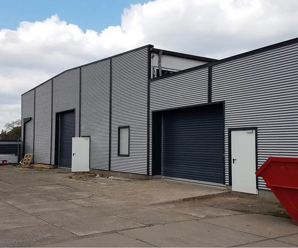 Industriehalle mit schwarz-weißen Streifen, großen Garagentoren und einem roten Müllcontainer auf einem Schotterplatz
