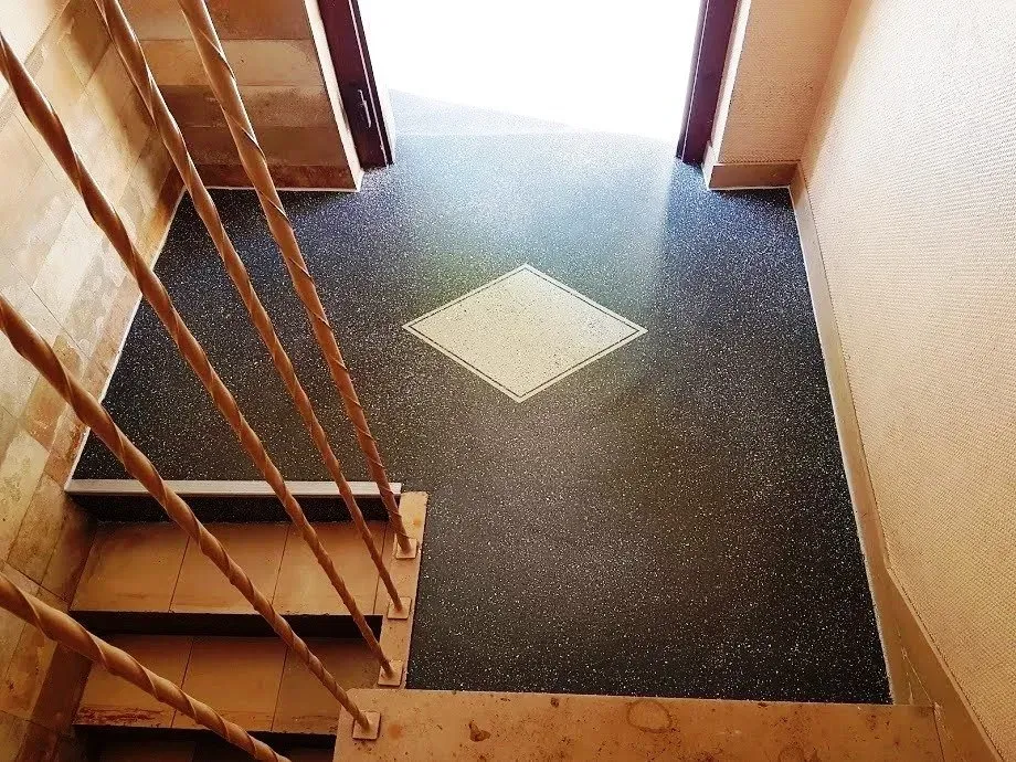 Treppenabsatz im Innenbereich mit dunkel gesprenkeltem Boden und einer quadratischen weißen Bodenfliese in der Nähe einer Tür