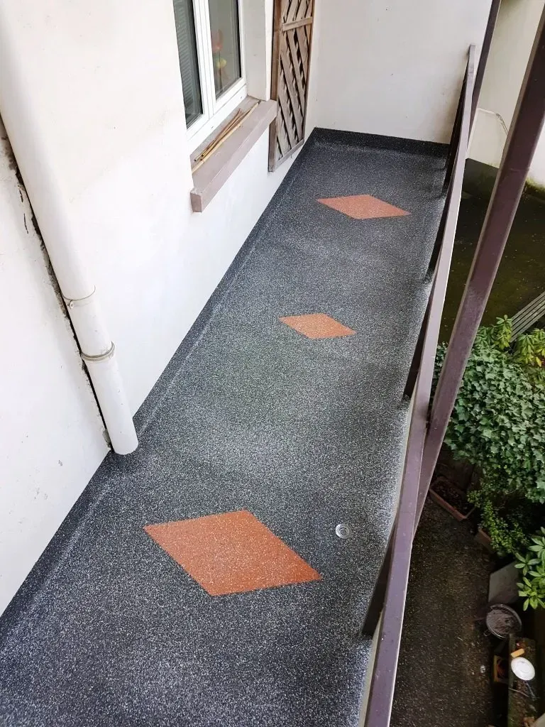 Schmaler Außenweg mit dunklem Teppich und orangefarbenen Rautenfliesen neben einer Hauswand und einem Gartengeländer