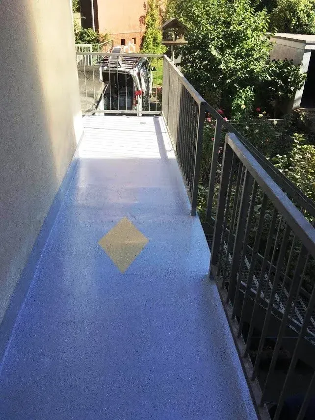 Ein blauer Gehweg mit gelber Rautenmarkierung neben einem schwarzen Geländer führt zu einem Außenpodest.