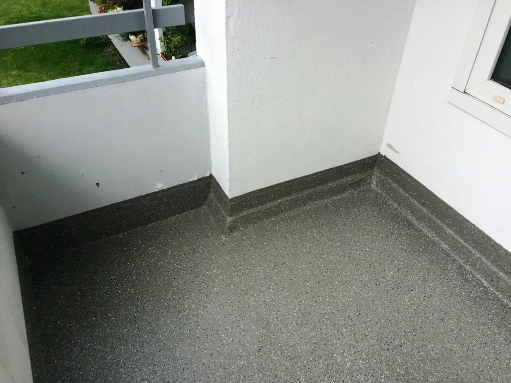 Kleine Balkonecke mit grauem Boden, weißen Wänden und einem Geländer mit Blick ins Grüne