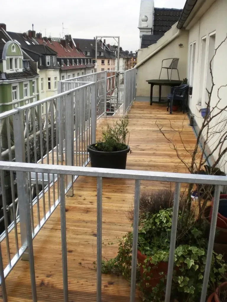 Schmaler Wohnungsbalkon mit Holzdielen, Metallgeländer, Topfpflanzen und einem kleinen Stuhl mit Blick auf die Gebäude
