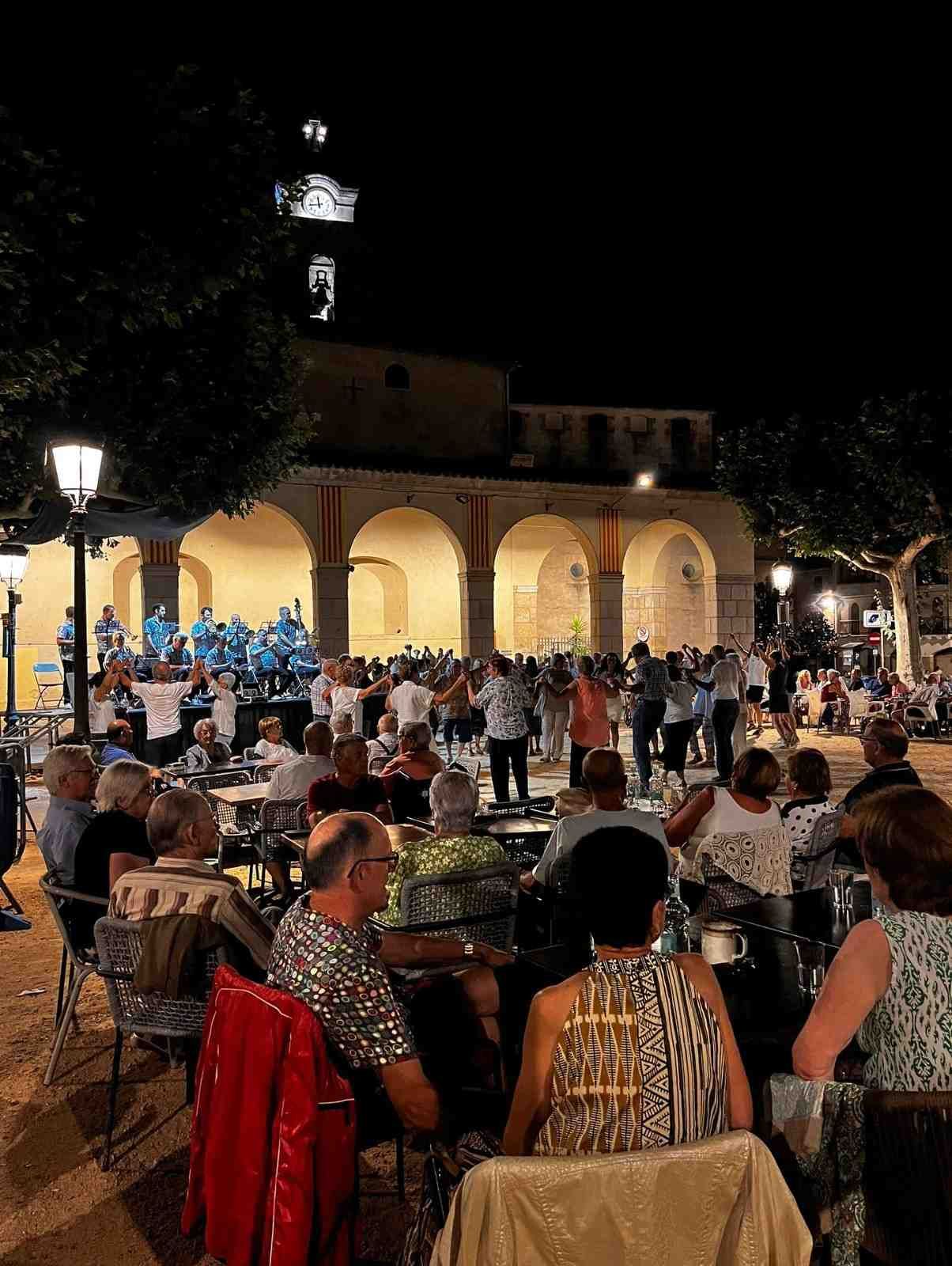 Plaza al aire libre abarrotada de gente por la noche, cenando en mesas junto a un edificio arqueado iluminado.