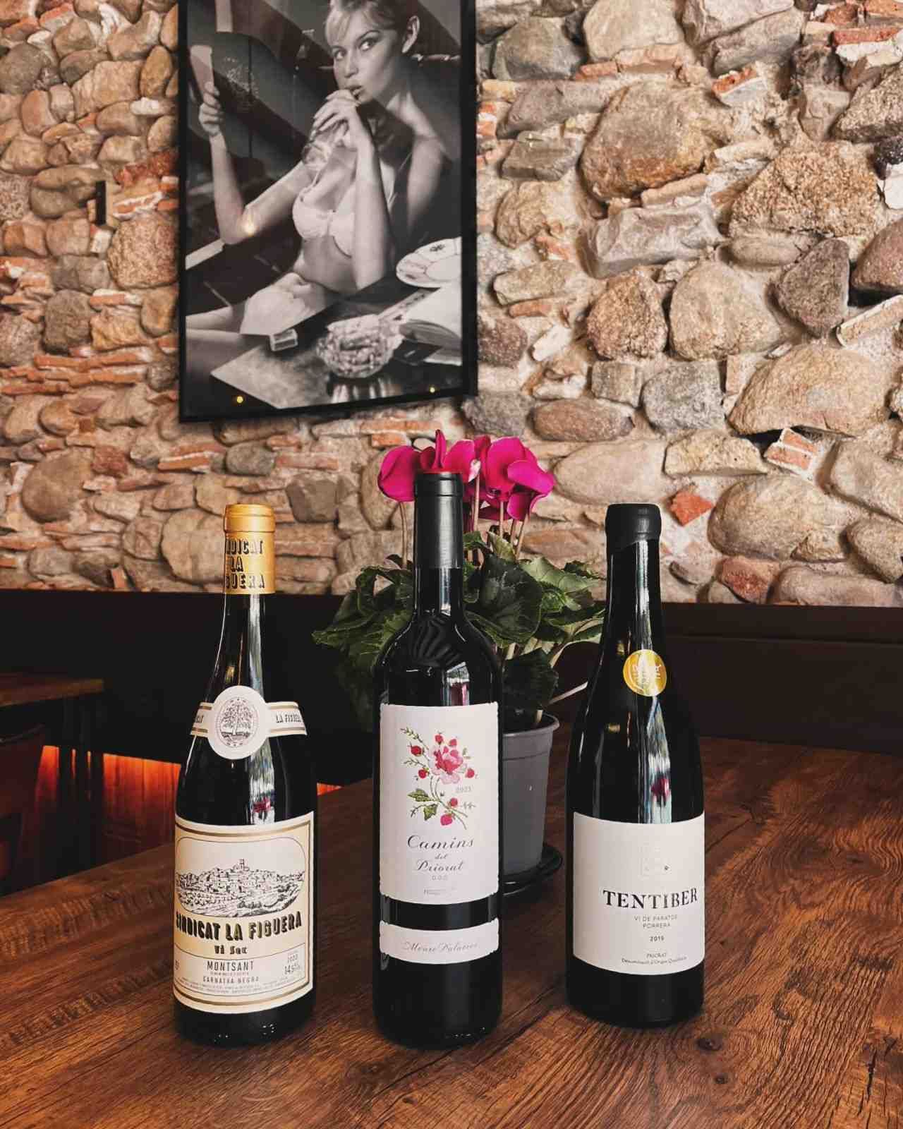 Tres botellas de vino sobre una mesa de madera frente a un muro de piedra con un retrato en blanco y negro.
