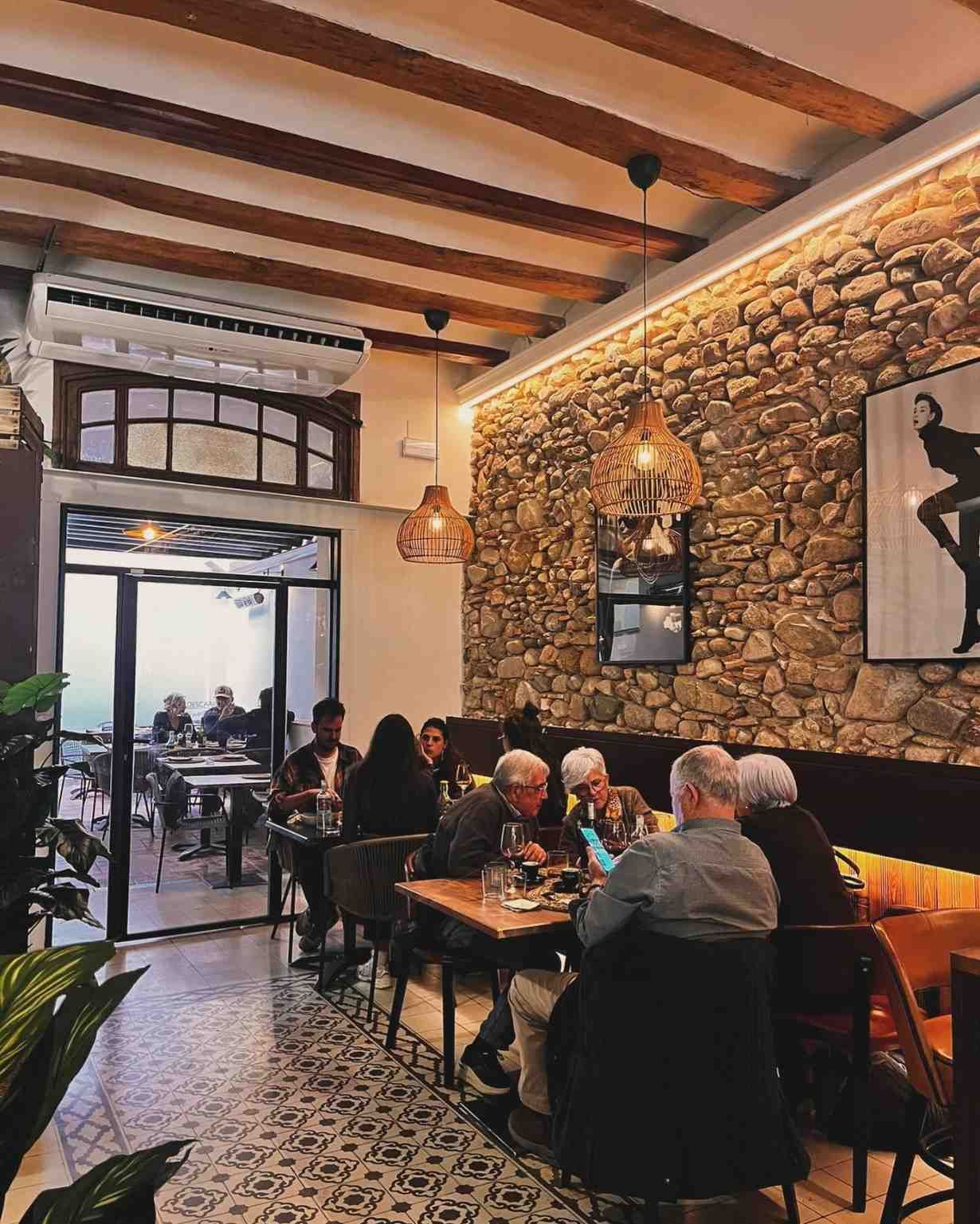Acogedora cafetería con paredes de piedra vista, suelo de baldosas estampadas y clientes sentados en mesas cerca de una gran puerta abierta.