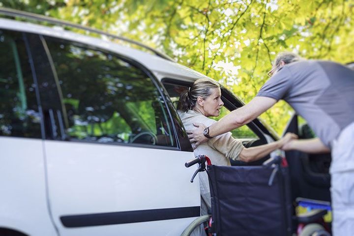 Une femme en fauteuil roulant est aidée à monter dans une voiture blanche par une autre personne, à l'extérieur, sous des arbres.