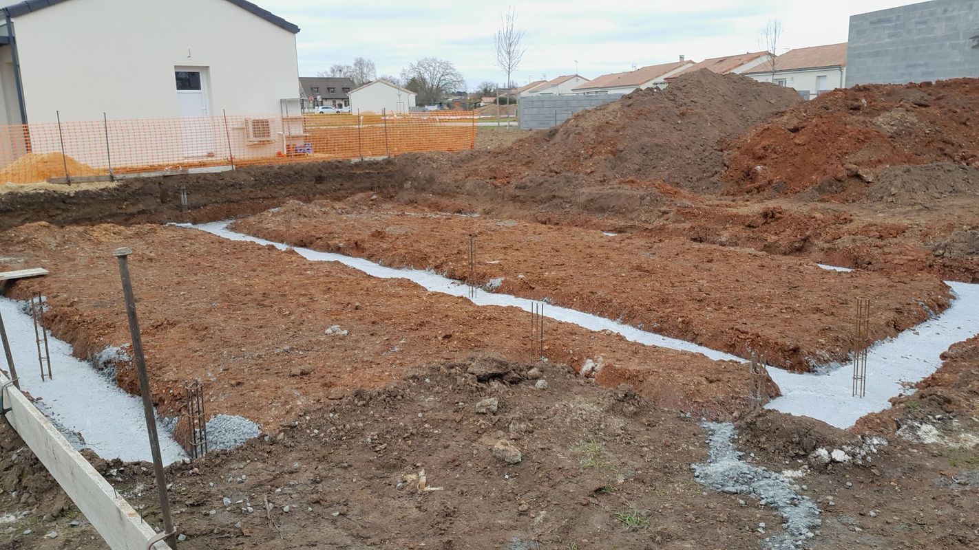 Chantier de construction avec des tas de terre excavée et des tranchées de fondation en béton à côté d'une maison