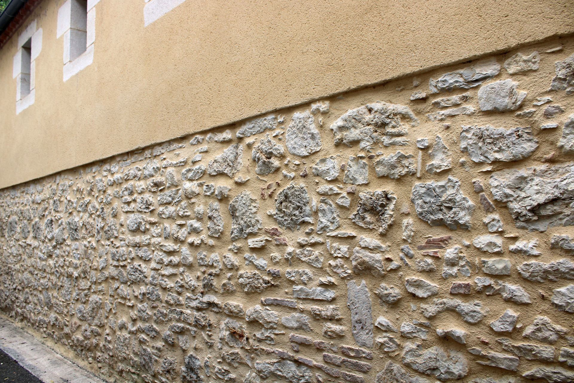 Mur de fondation en pierre sous un revêtement extérieur en stuc beige d'un bâtiment, vu en biais.