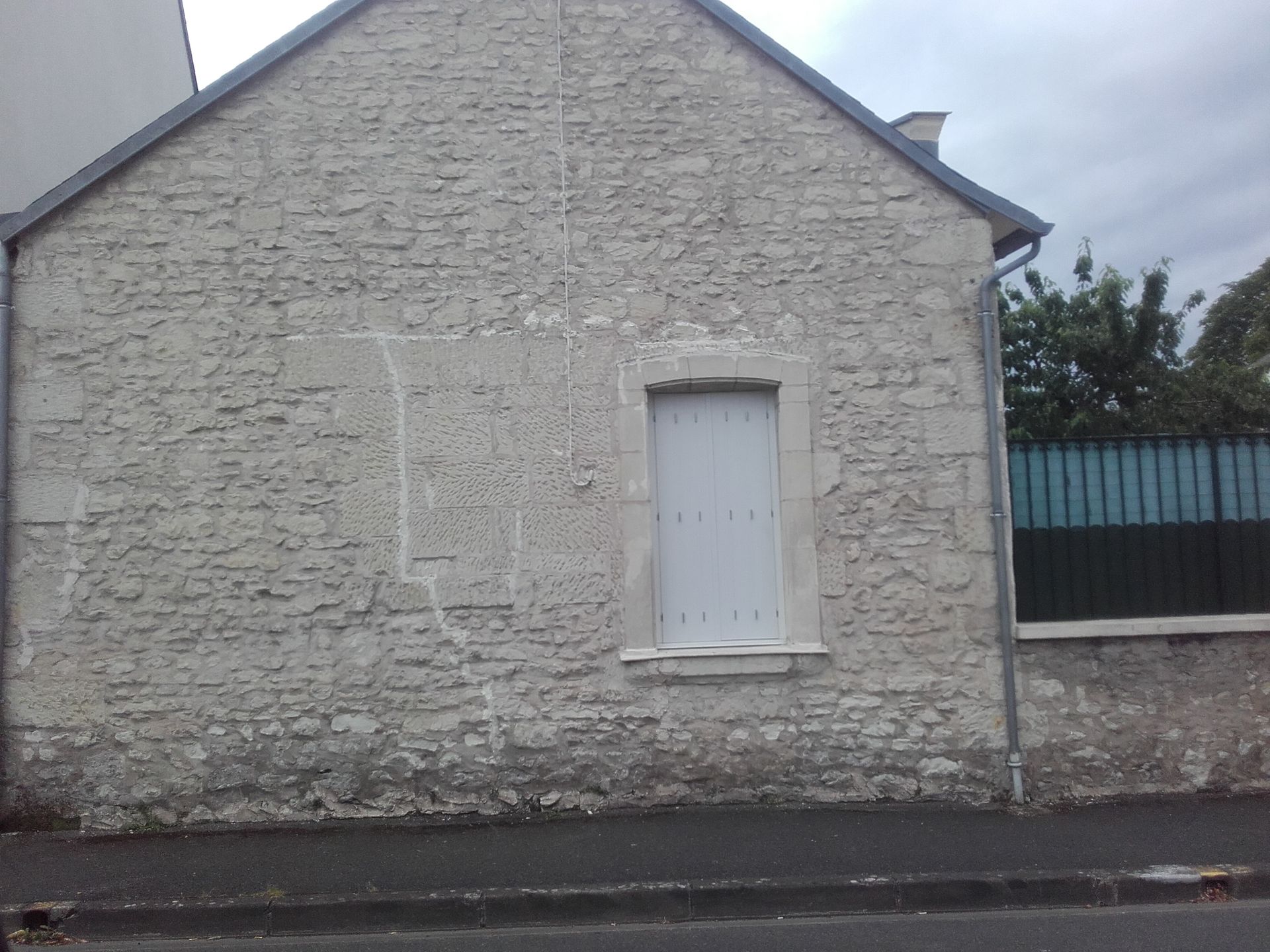 Mur en pierre d'une maison avec une petite fenêtre à volets blancs donnant sur une rue