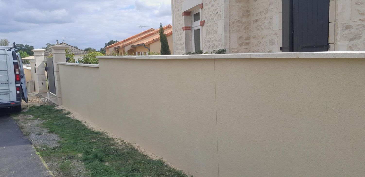 Un mur beige borde une rue étroite, à côté d'une camionnette blanche garée et de maisons sous un ciel nuageux.