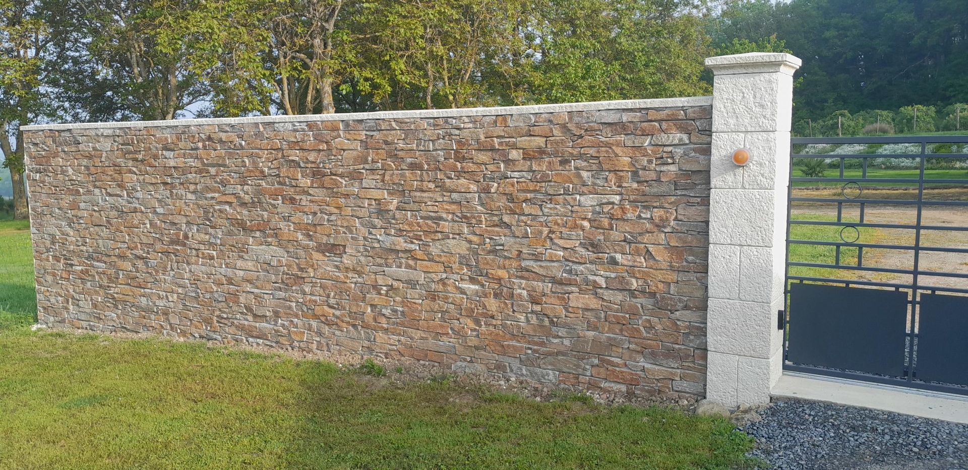 Un mur de pierre avec un pilier blanc à côté d'un portail en métal noir.