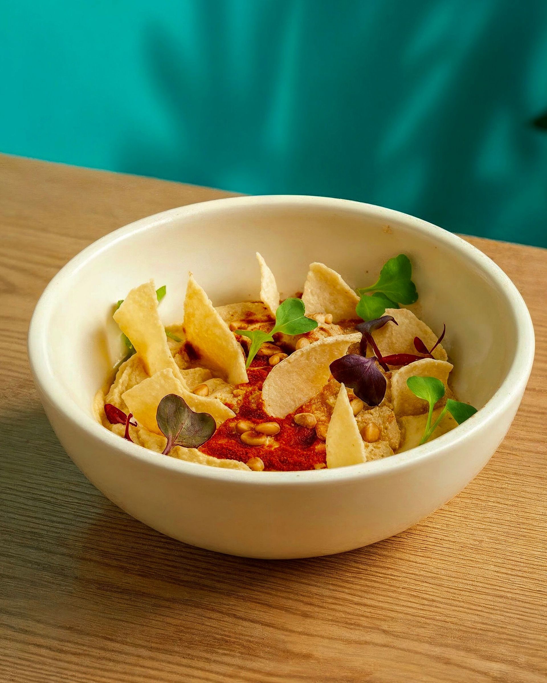 Tazón blanco de hummus cubierto con salsa roja, patatas fritas y hierbas sobre una mesa de madera.