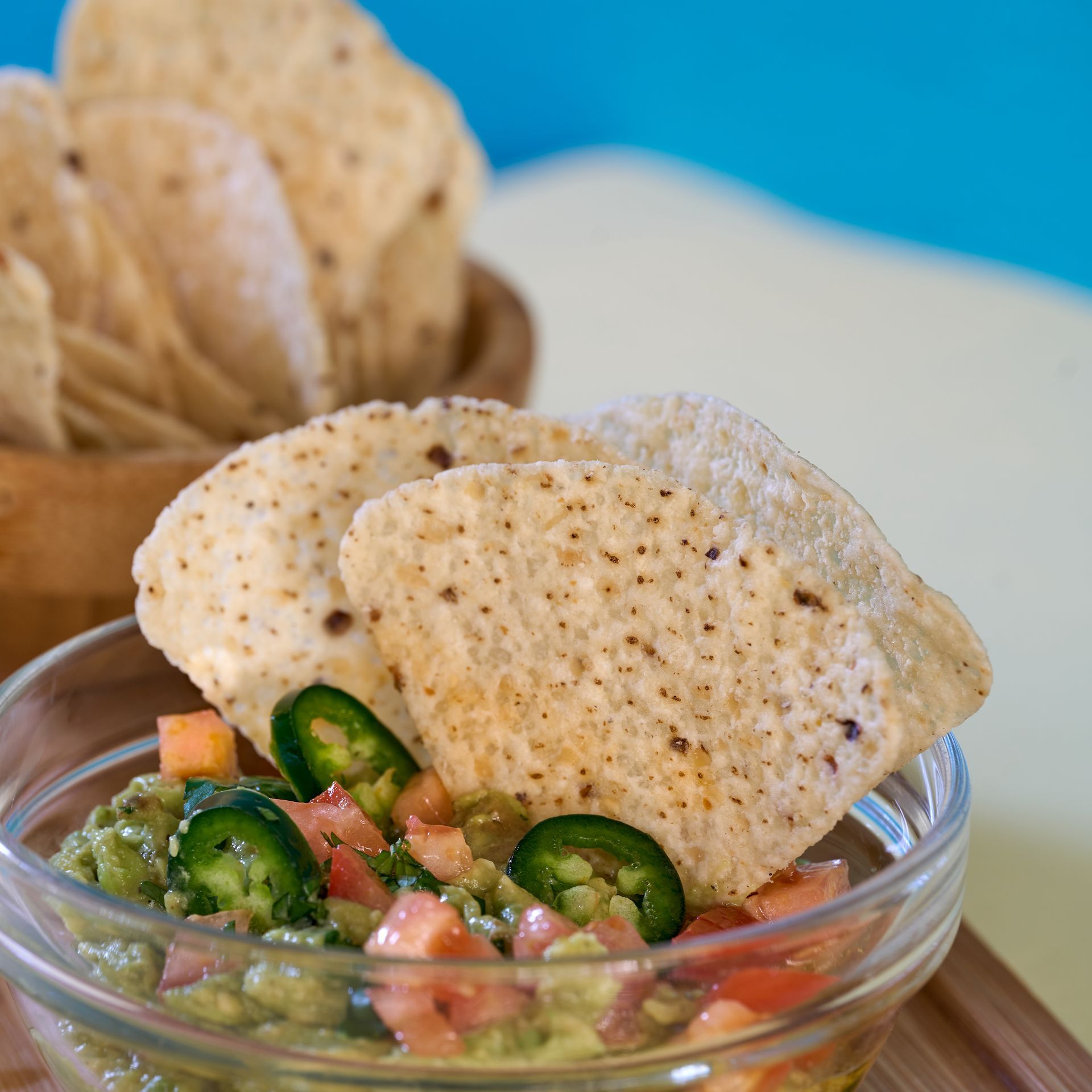 Guacamole con totopos en un recipiente de vidrio, con un recipiente de madera con totopos al fondo.