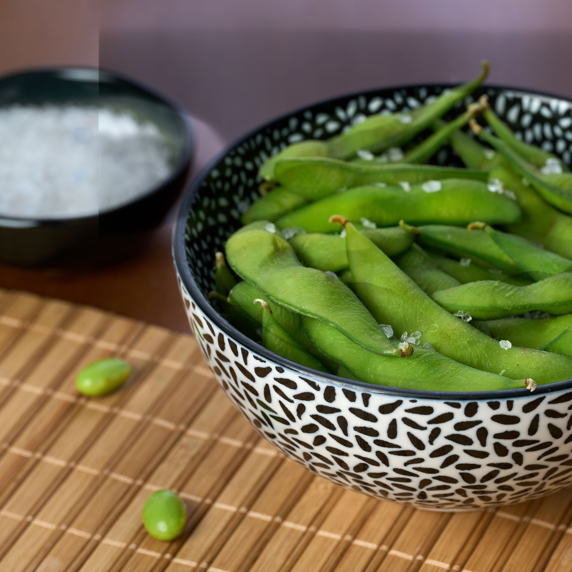 Cuenco de edamame con sal, sobre una estera de bambú, con un pequeño cuenco de sal cerca.