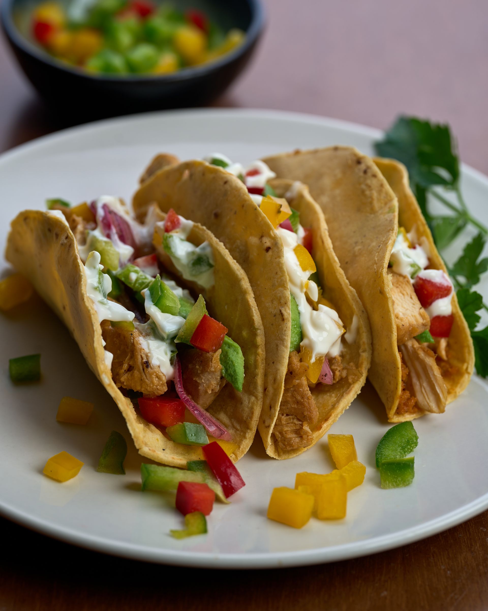 Tres tacos en un plato blanco, rellenos de pollo, pimientos y crema, con un tazón de salsa de fondo.