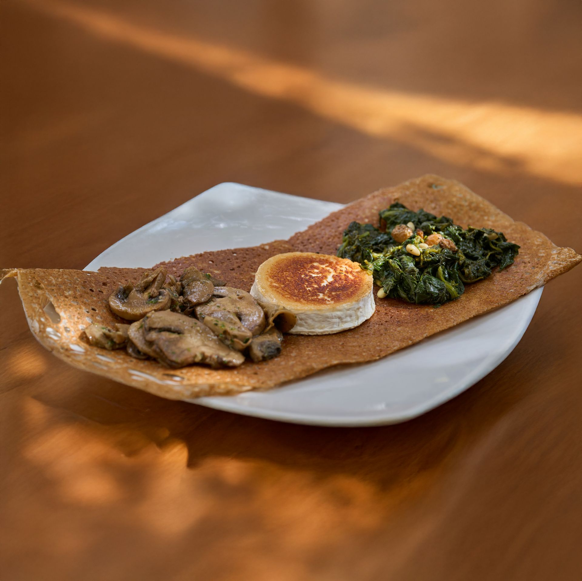 Crepe sabrosa de trigo sarraceno con champiñones, espinacas y una rodaja de queso de cabra en un plato blanco.