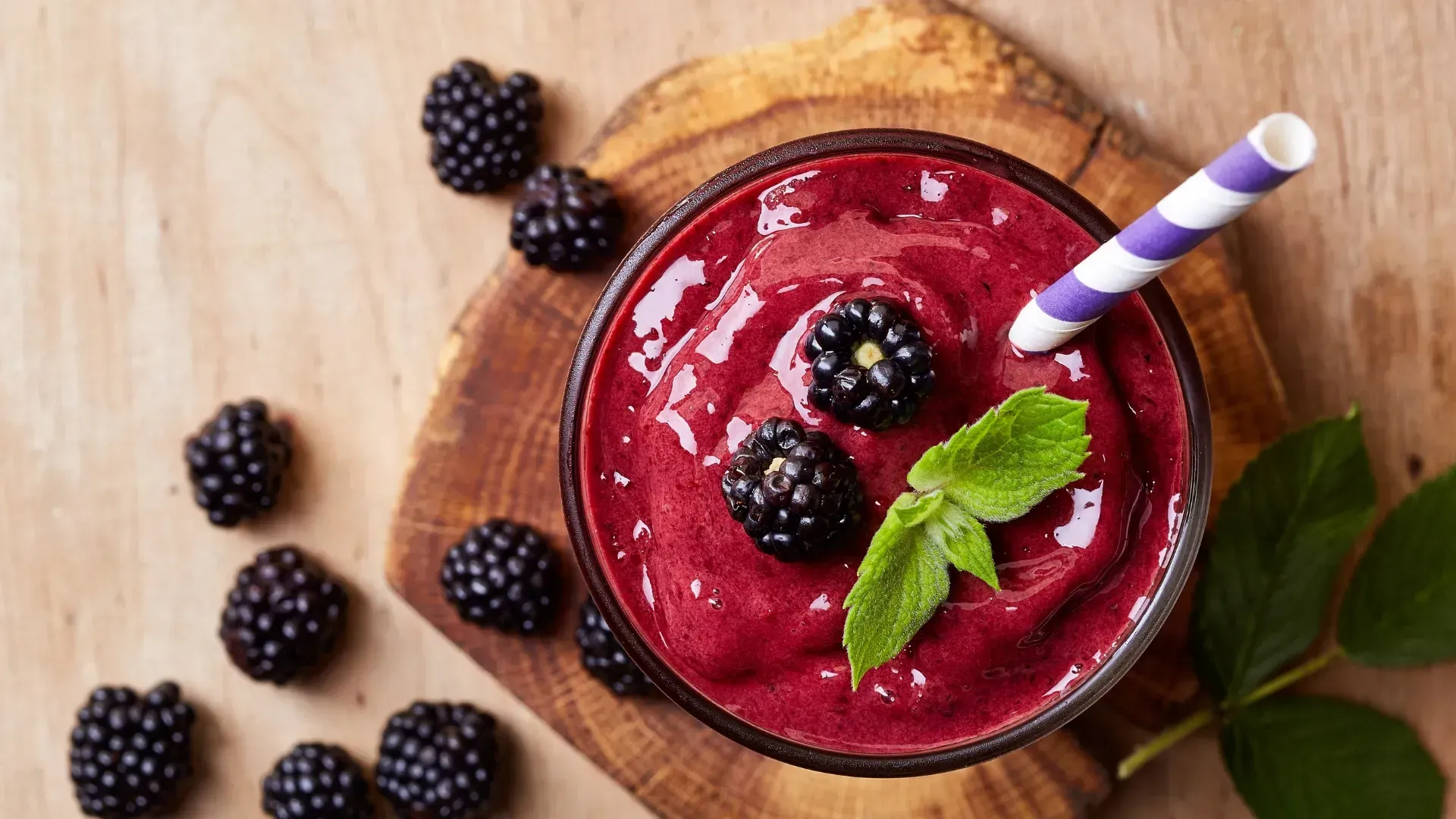 Batido de moras en vaso con pajita, adornado con moras y menta, sobre una tabla de madera con moras esparcidas.