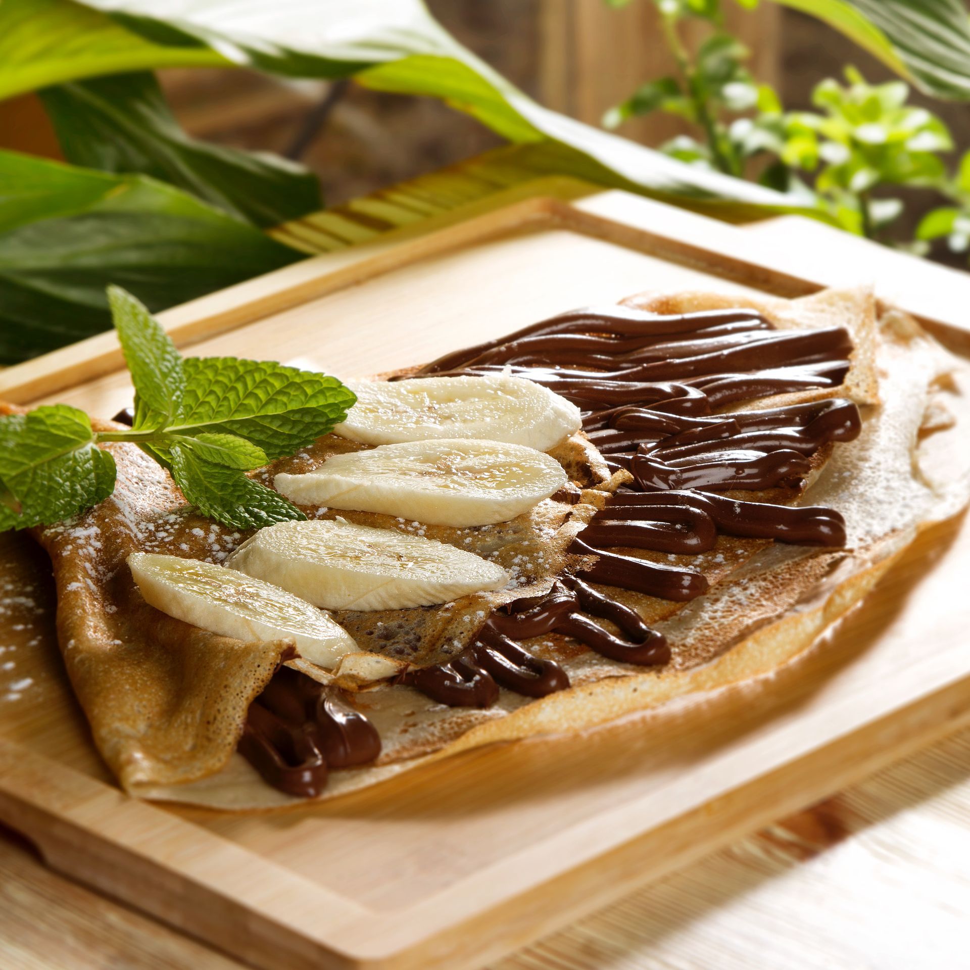 Crepe con rodajas de plátano y llovizna de chocolate sobre una tabla de madera, adornado con menta.