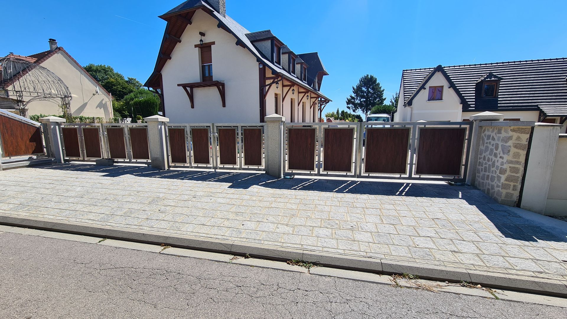 Maison d'habitation aux murs blancs et au portail marron, située le long d'une rue pavée sous un ciel d'un bleu limpide.