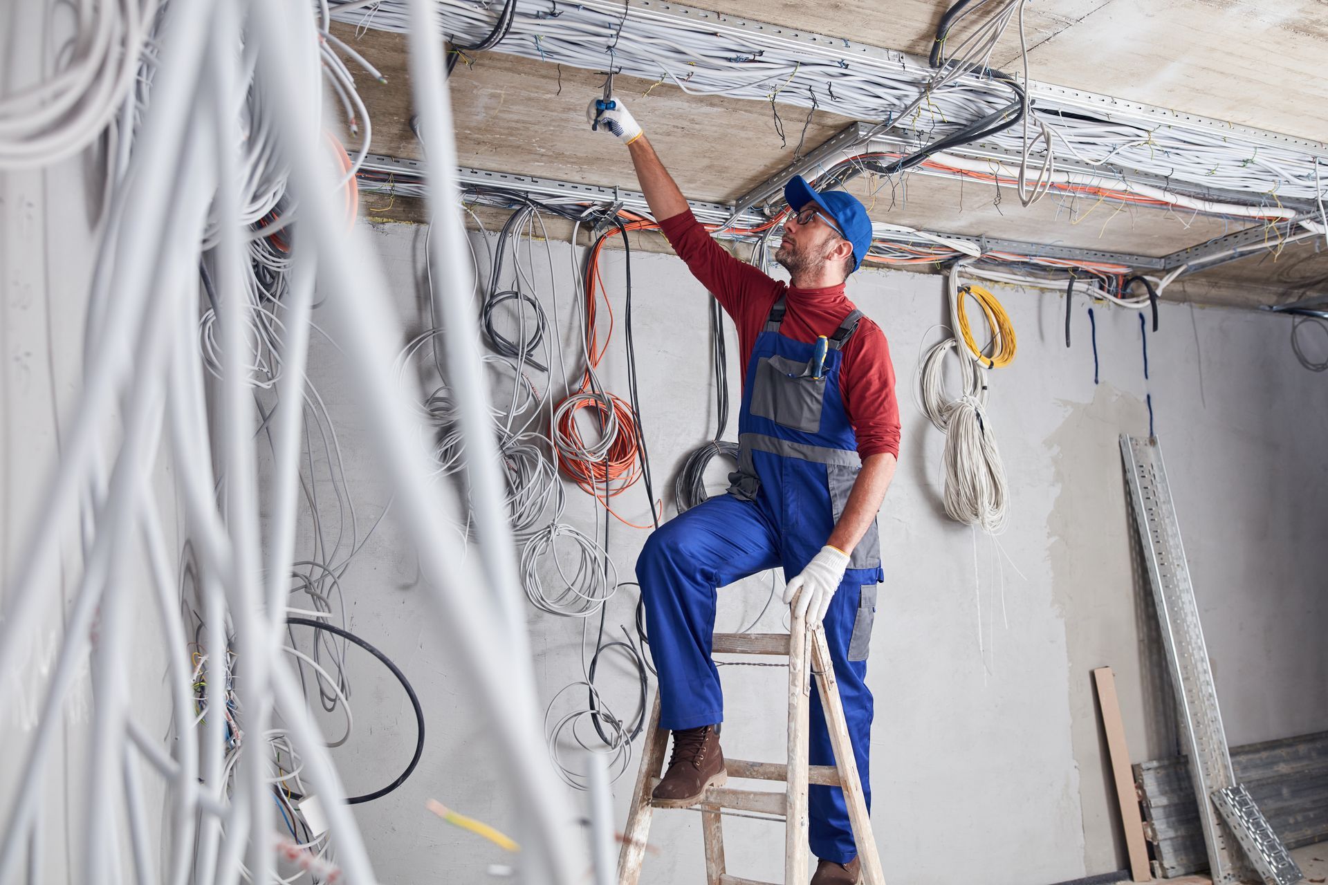 Sur un chantier, un ouvrier en salopette bleue effectue des travaux de câblage au plafond, des câbles et des outils apparents se trouvant autour de lui.