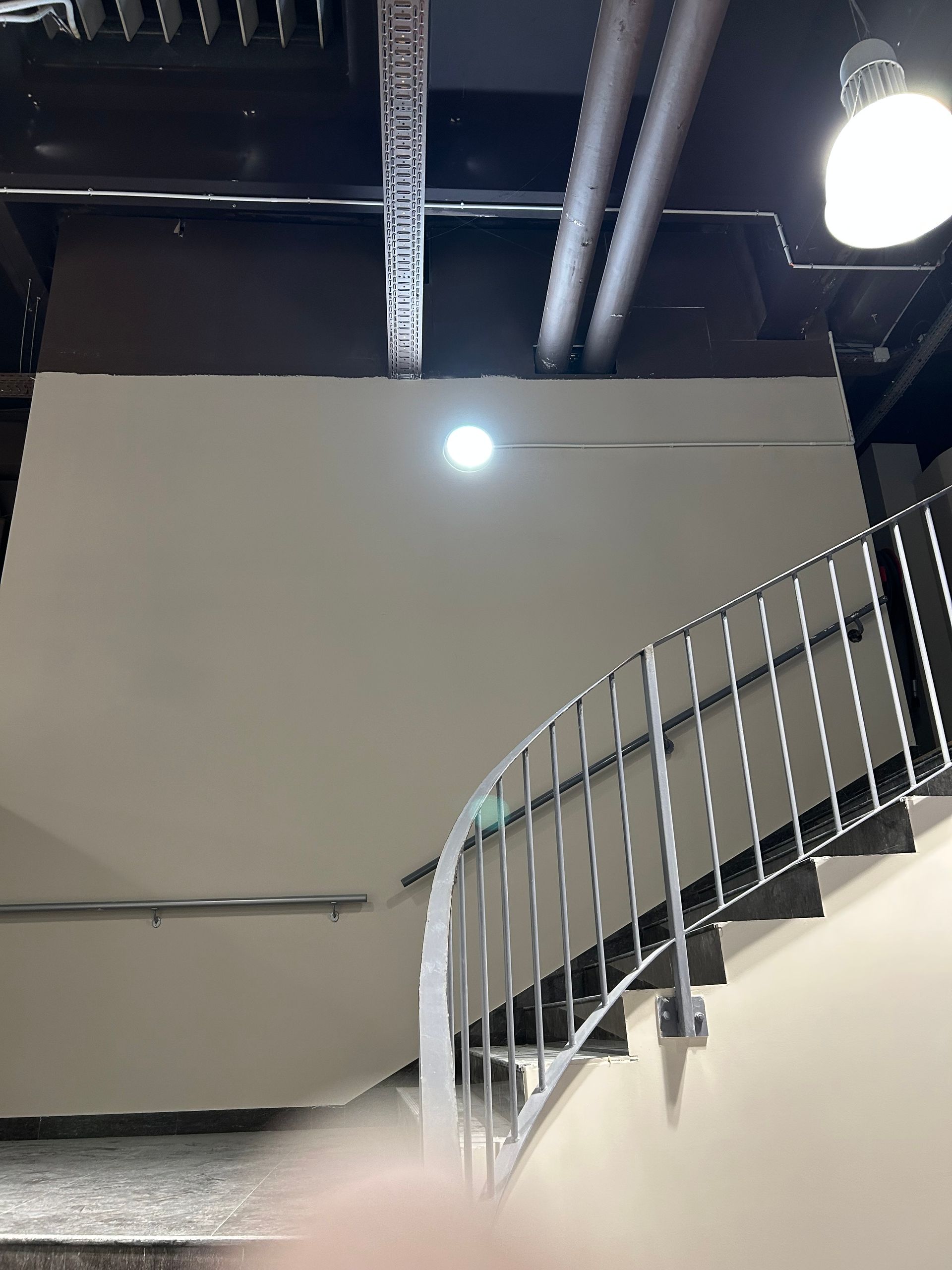 Escalier métallique incurvé avec rampe blanche dans un intérieur industriel sous des tuyaux et des luminaires apparents au plafond.