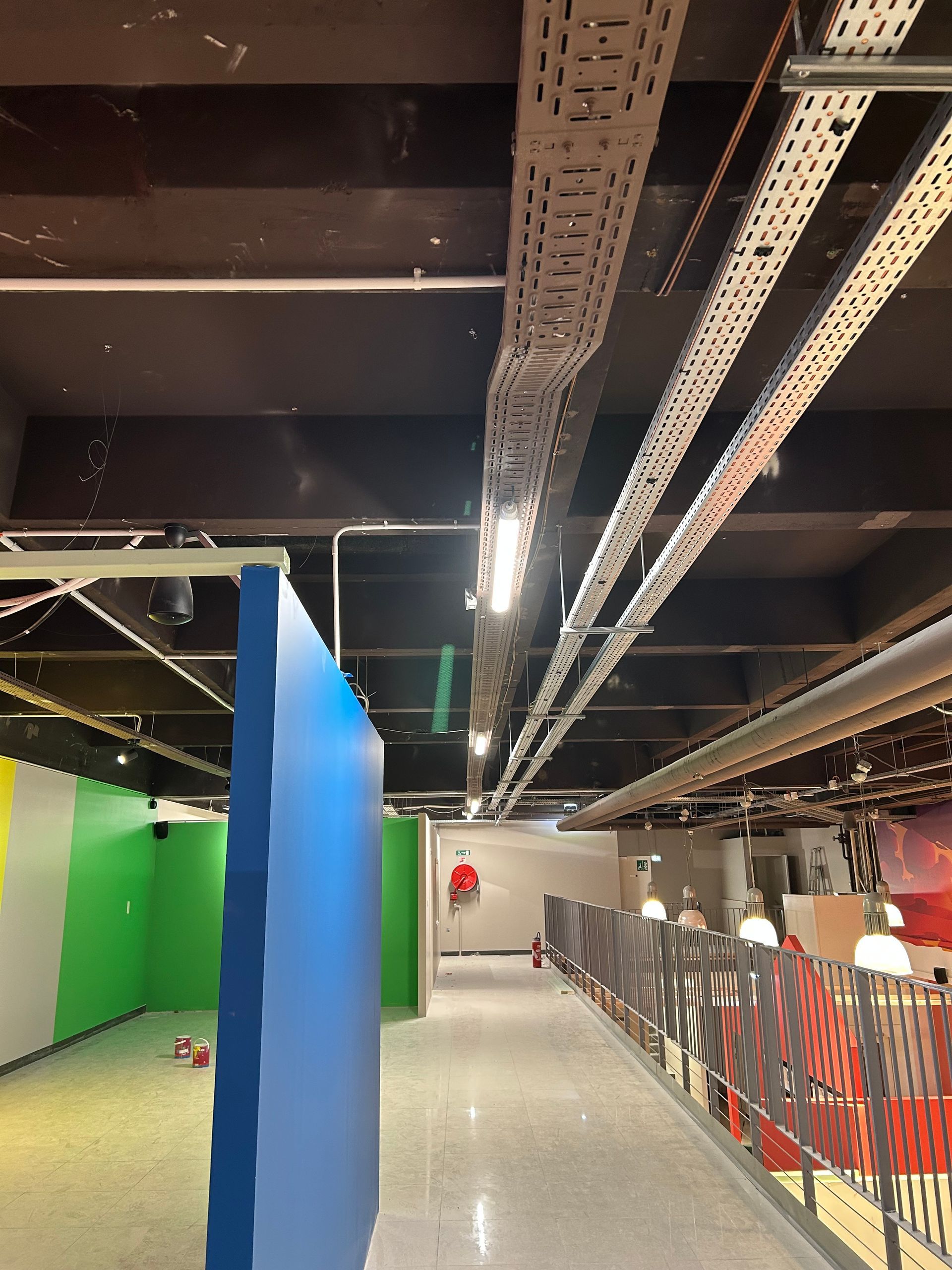 Couloir intérieur avec pilier bleu, poutres apparentes au plafond et panneaux muraux verts à côté d'une rambarde métallique.