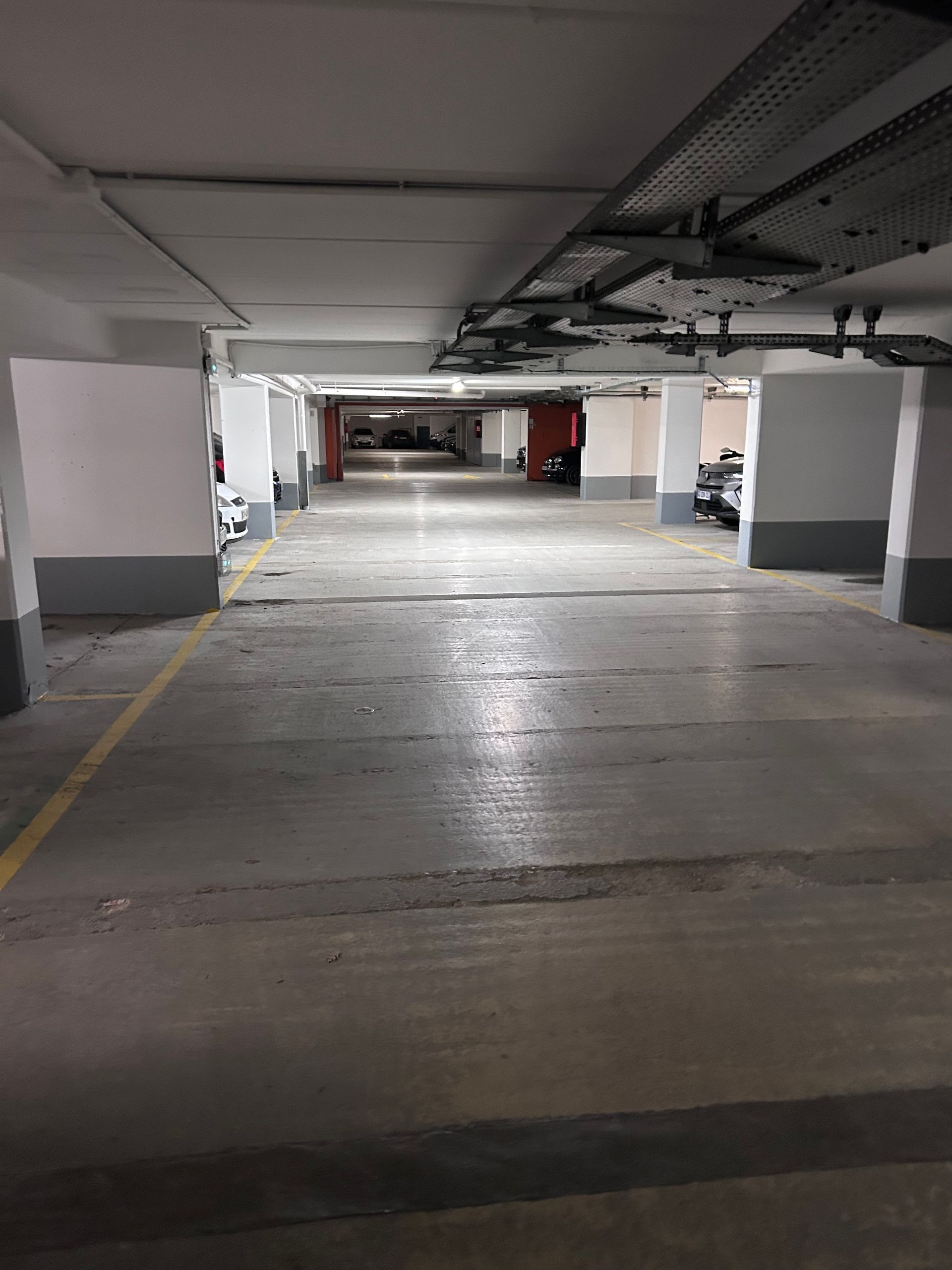 Parking souterrain vide avec piliers en béton et tuyaux au plafond, éclairé par des néons.